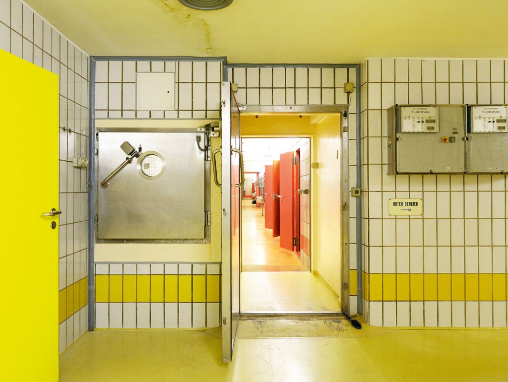 Ein gelber Raum mit Fliesen, mit Blick auf einen Gang mit roten Türen zu kleineren Zellen und Tierställen